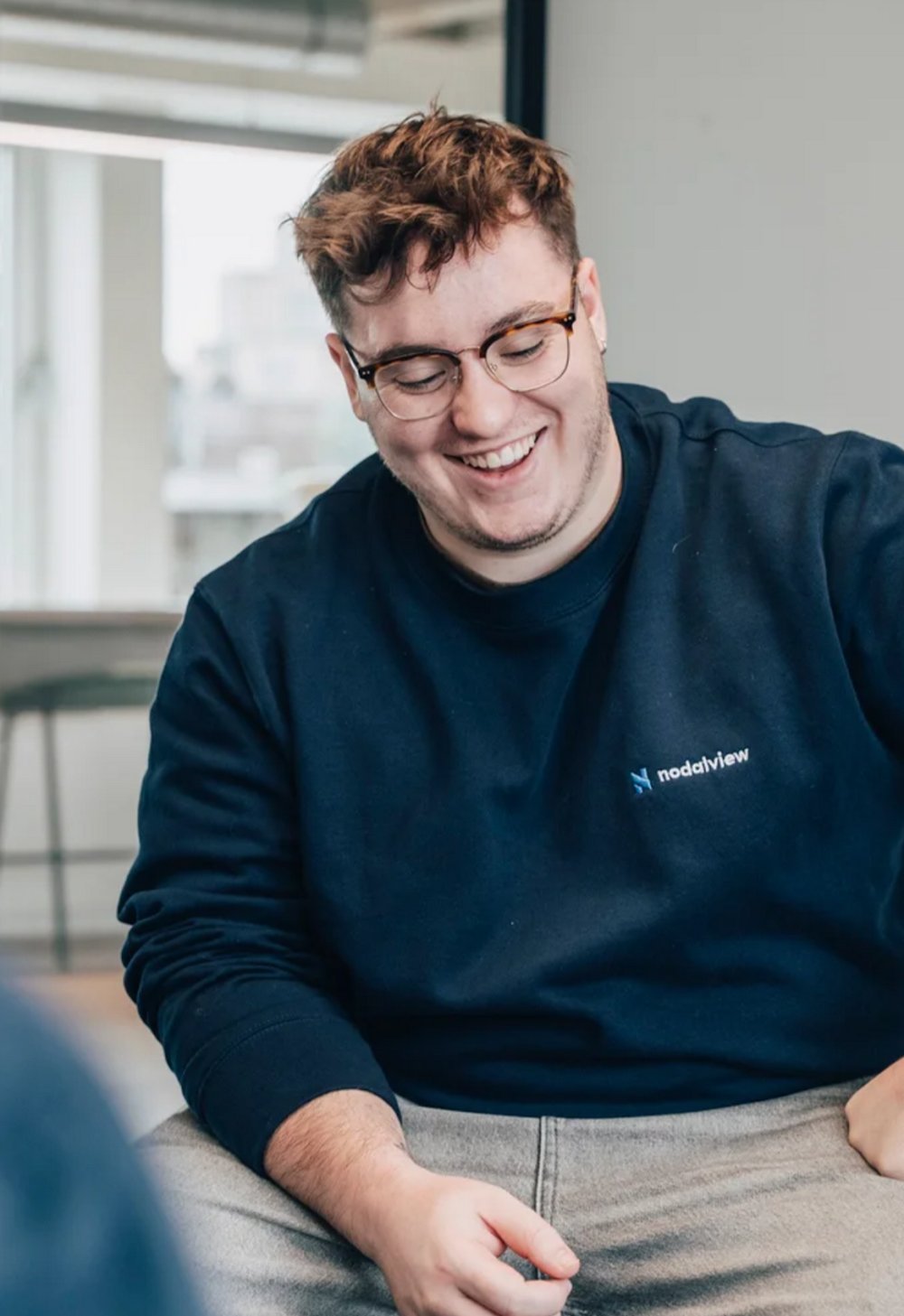 Aymeric Carlier in a dark Nodalview sweatshirt and glasses, laughing, in an office by a window.